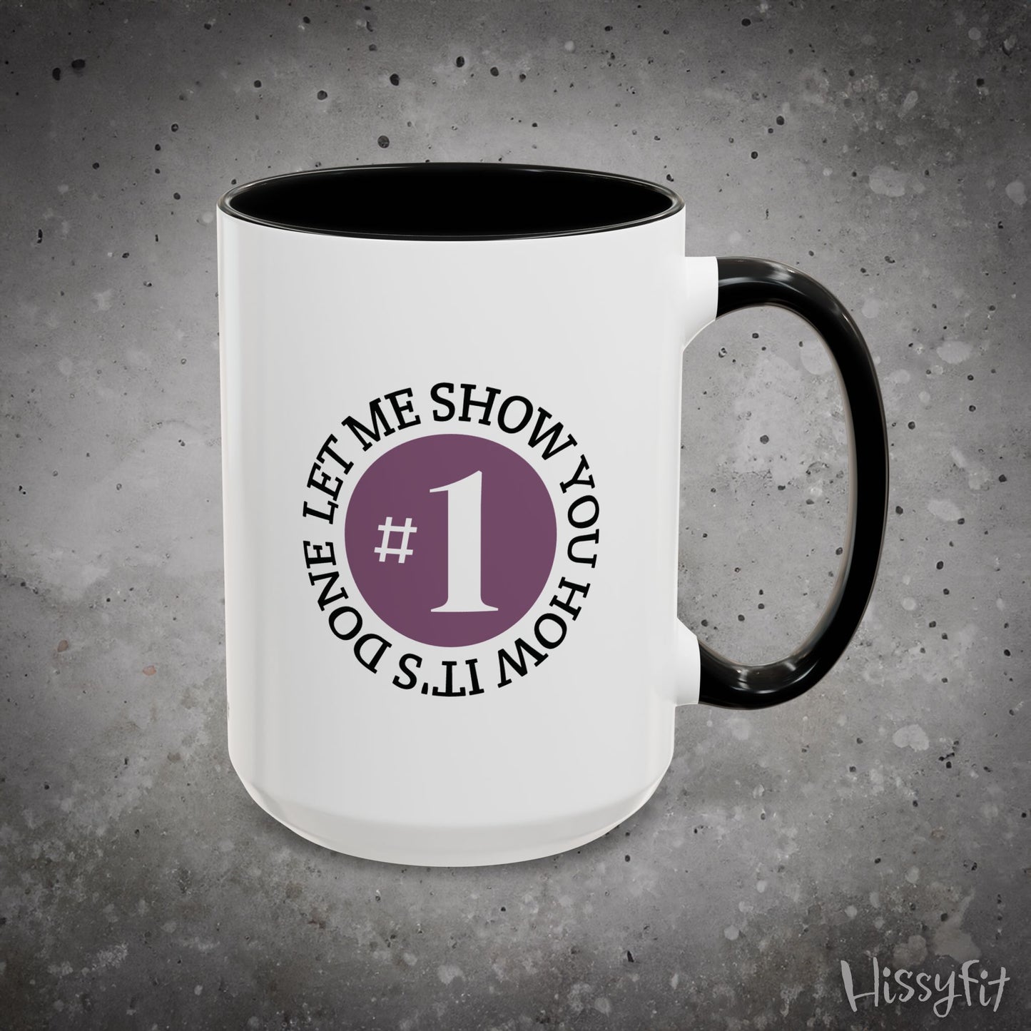 White mug with black interior and handle on a gray background, featuring text 'Let me show you how it's done' and a number one symbol.