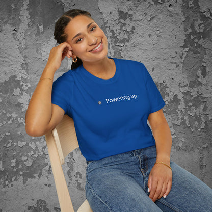 Woman wearing a blue t-shirt with 'Powering up' text, sitting against a textured gray wall.
