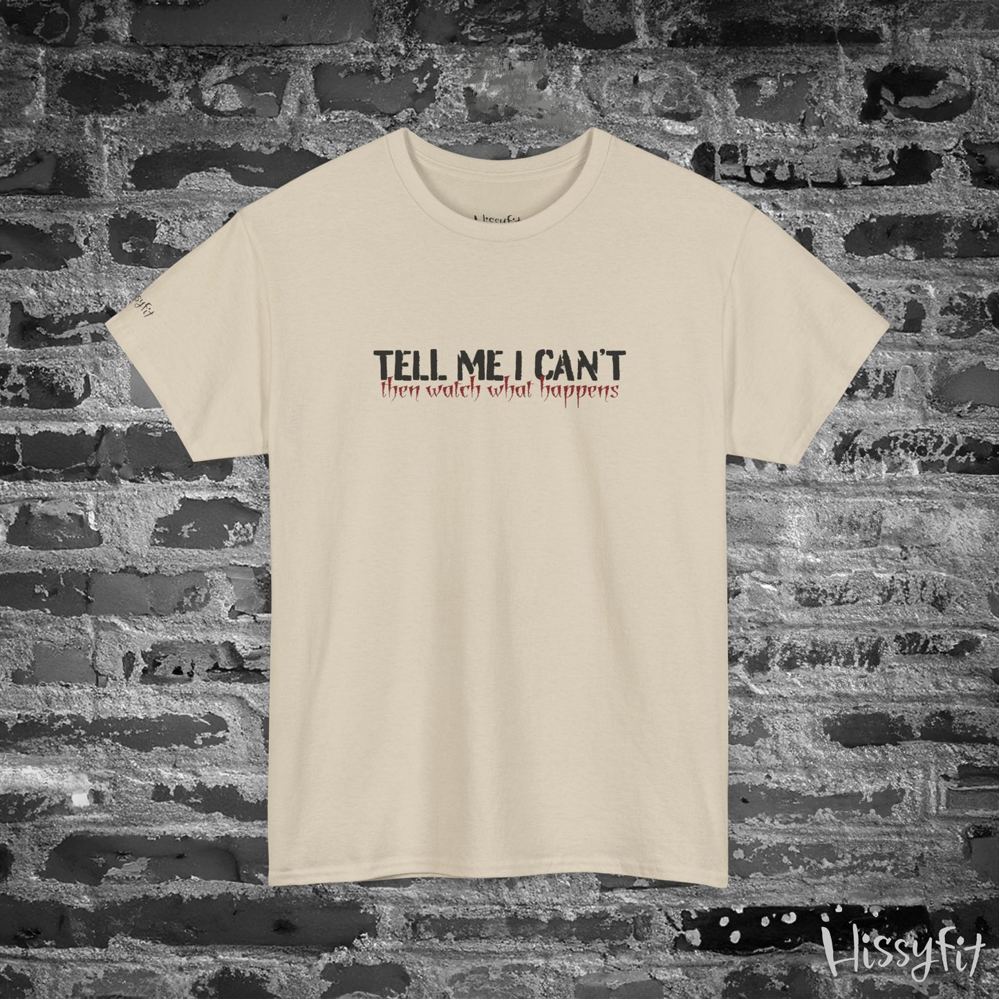 Beige t-shirt with text 'Tell me I can't then watch what happens' on a brick wall background
