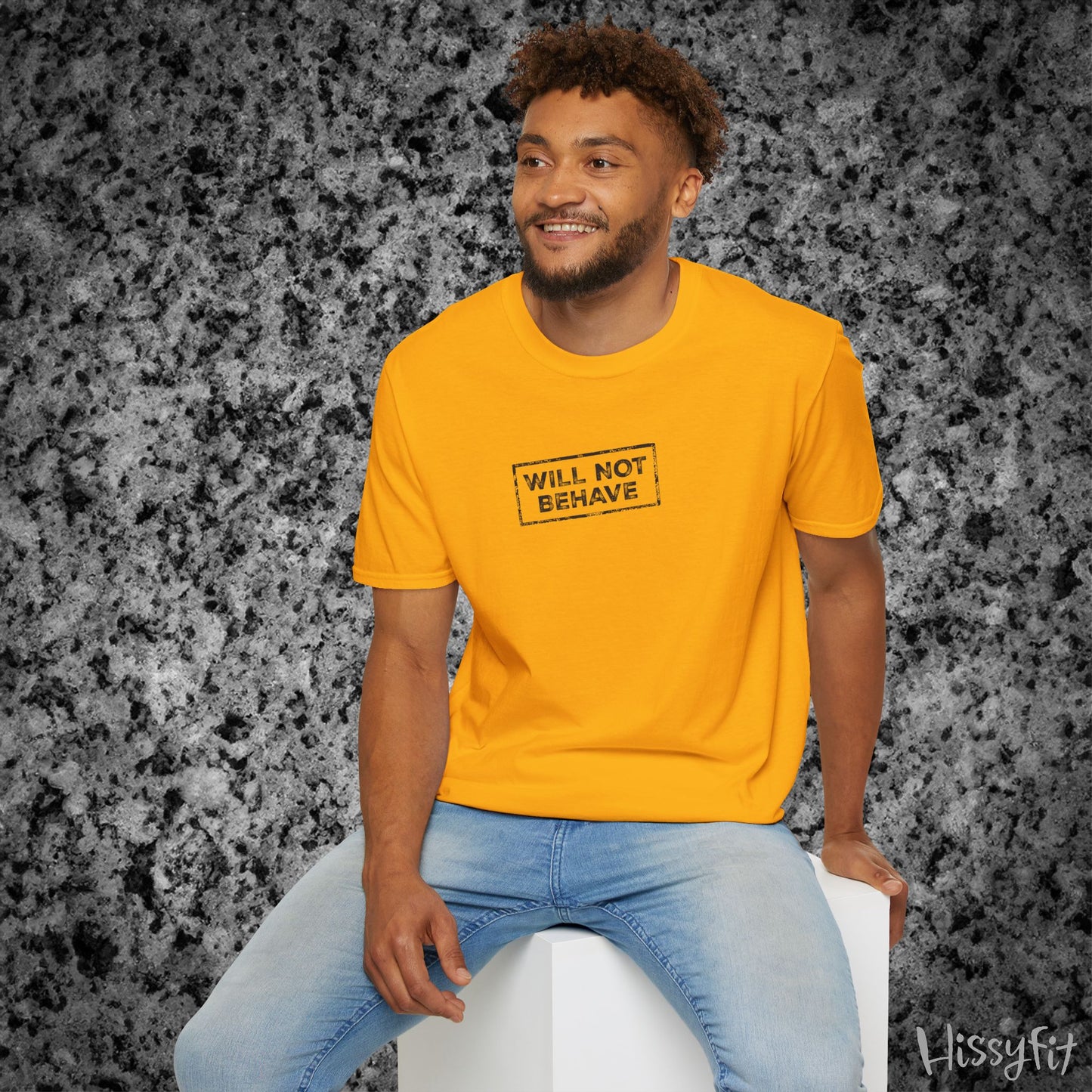 Man wearing a yellow t-shirt with 'WILL NOT BEHAVE' text, sitting on a white stool against a gray textured background.