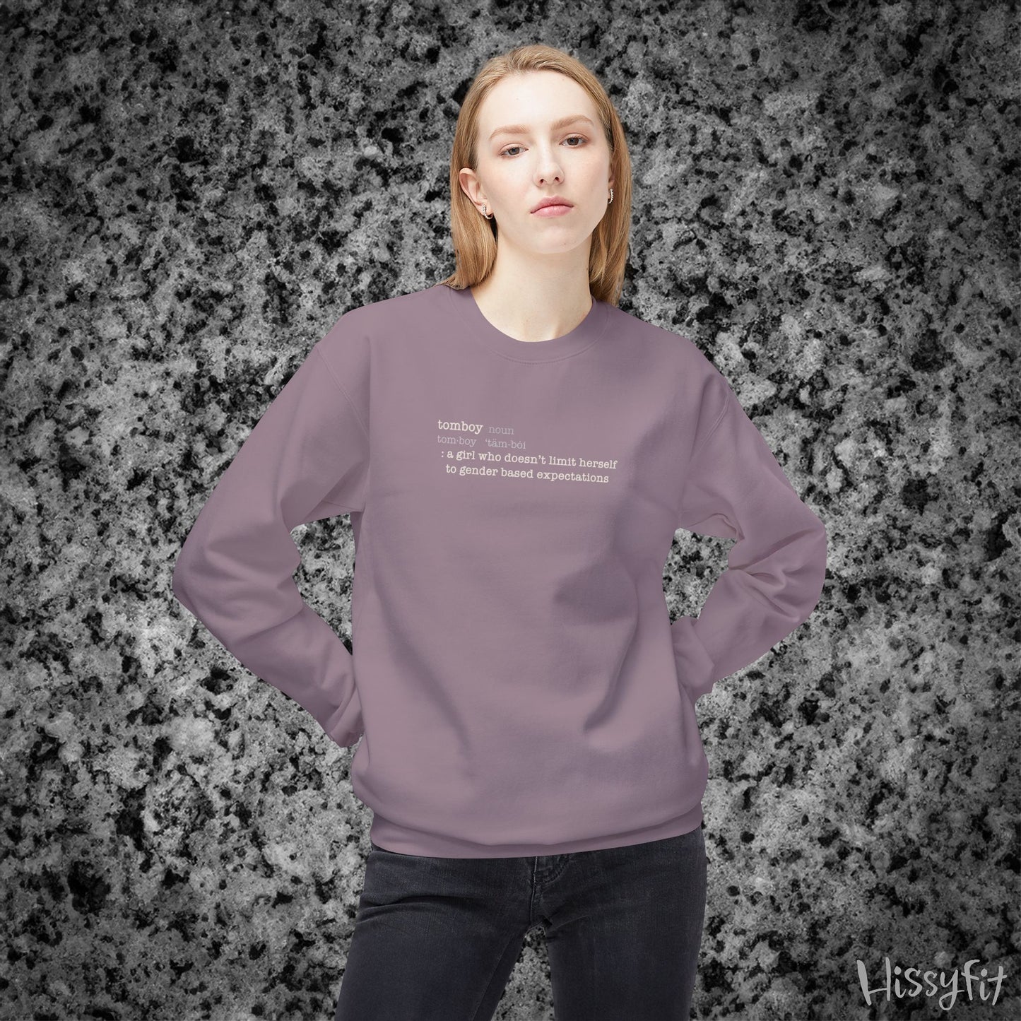 Person wearing a light purple sweatshirt with text definition of "Tomboy" on a gray and black speckled background