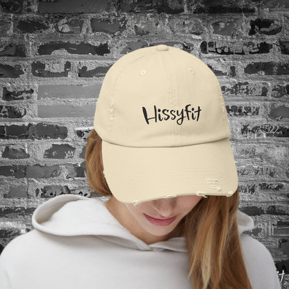 Person wearing a beige cap with 'Hissyfit' text against a textured wall background