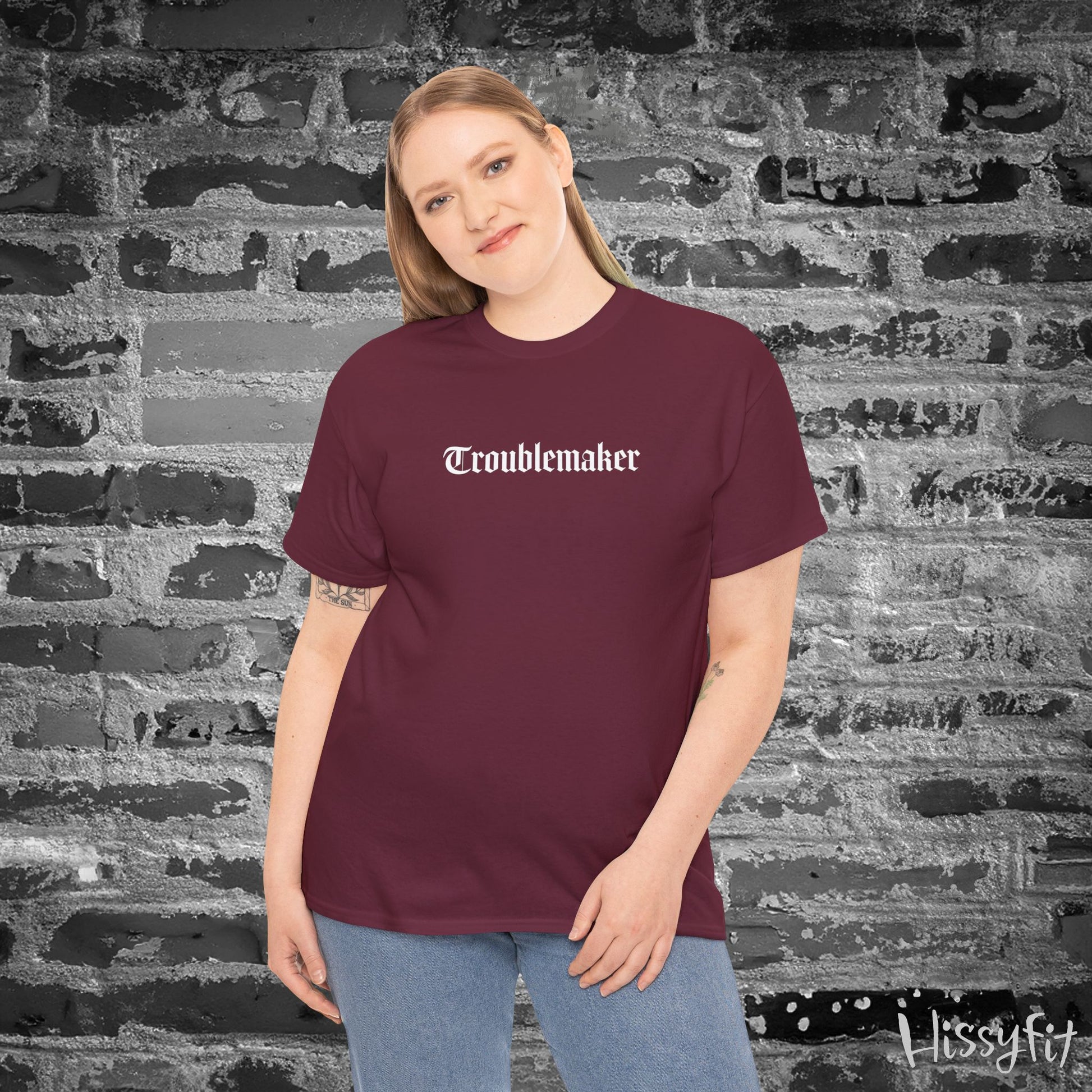Person wearing a maroon t-shirt with 'Troublemaker' text against a textured wall.
