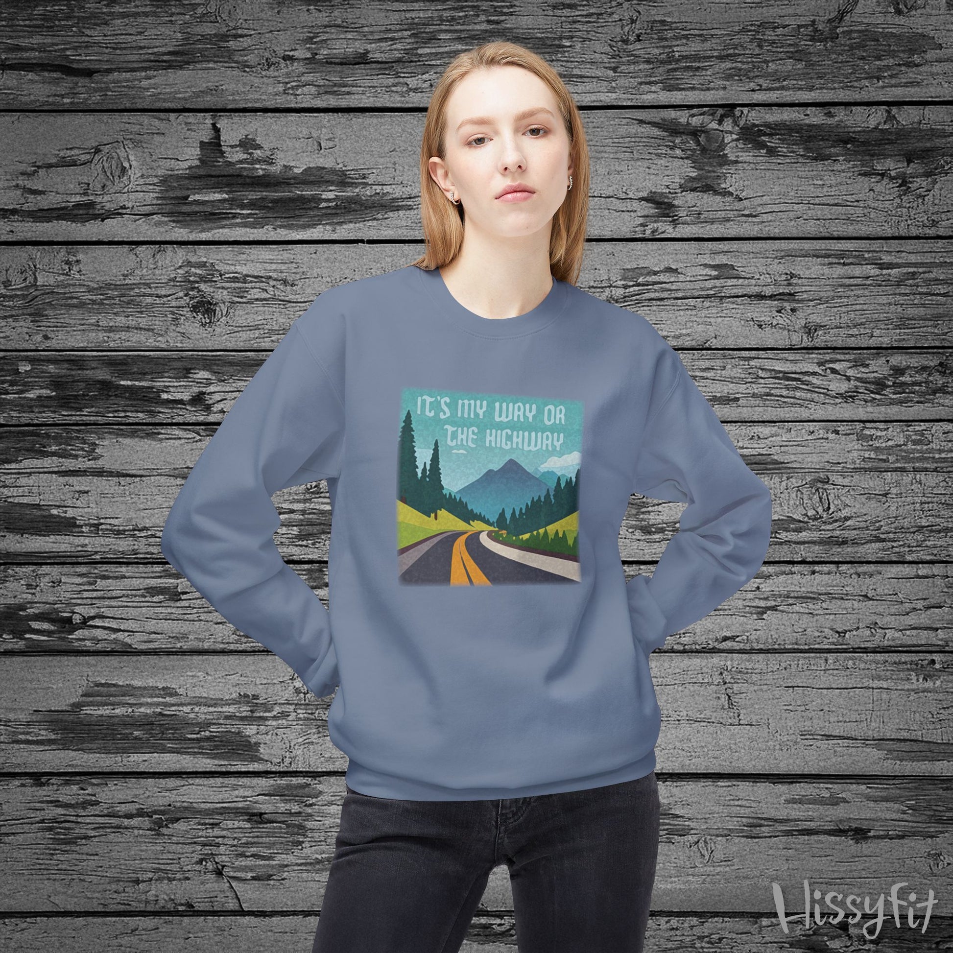 Person wearing a blue sweatshirt with a graphic design of a road and mountains and "It's my way or the highway" text on a wooden background