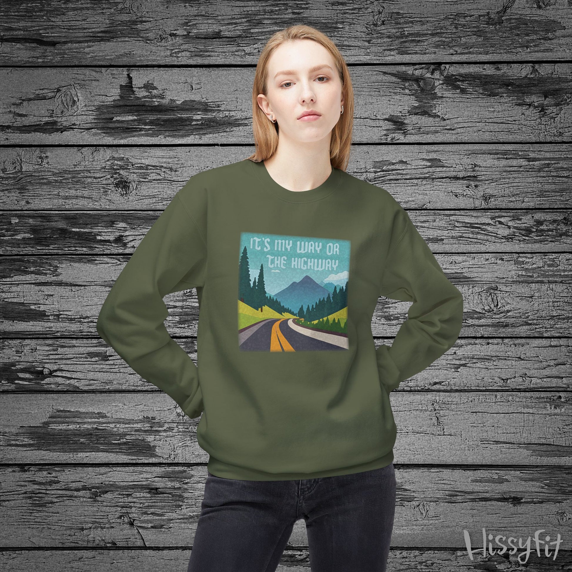 Person wearing a green sweatshirt with a graphic design of a road and mountains and "It's my way or the highway" text on a wooden background