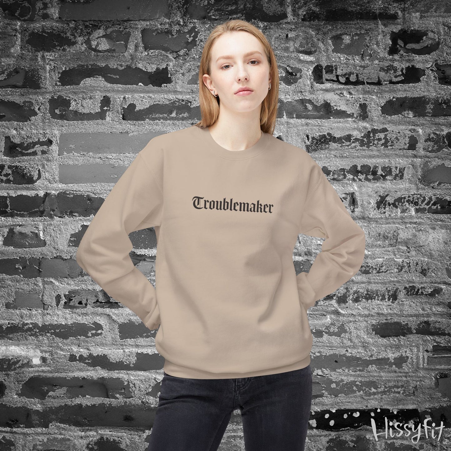 Person wearing a beige sweatshirt with 'Troublemaker' text against a textured brick wall.