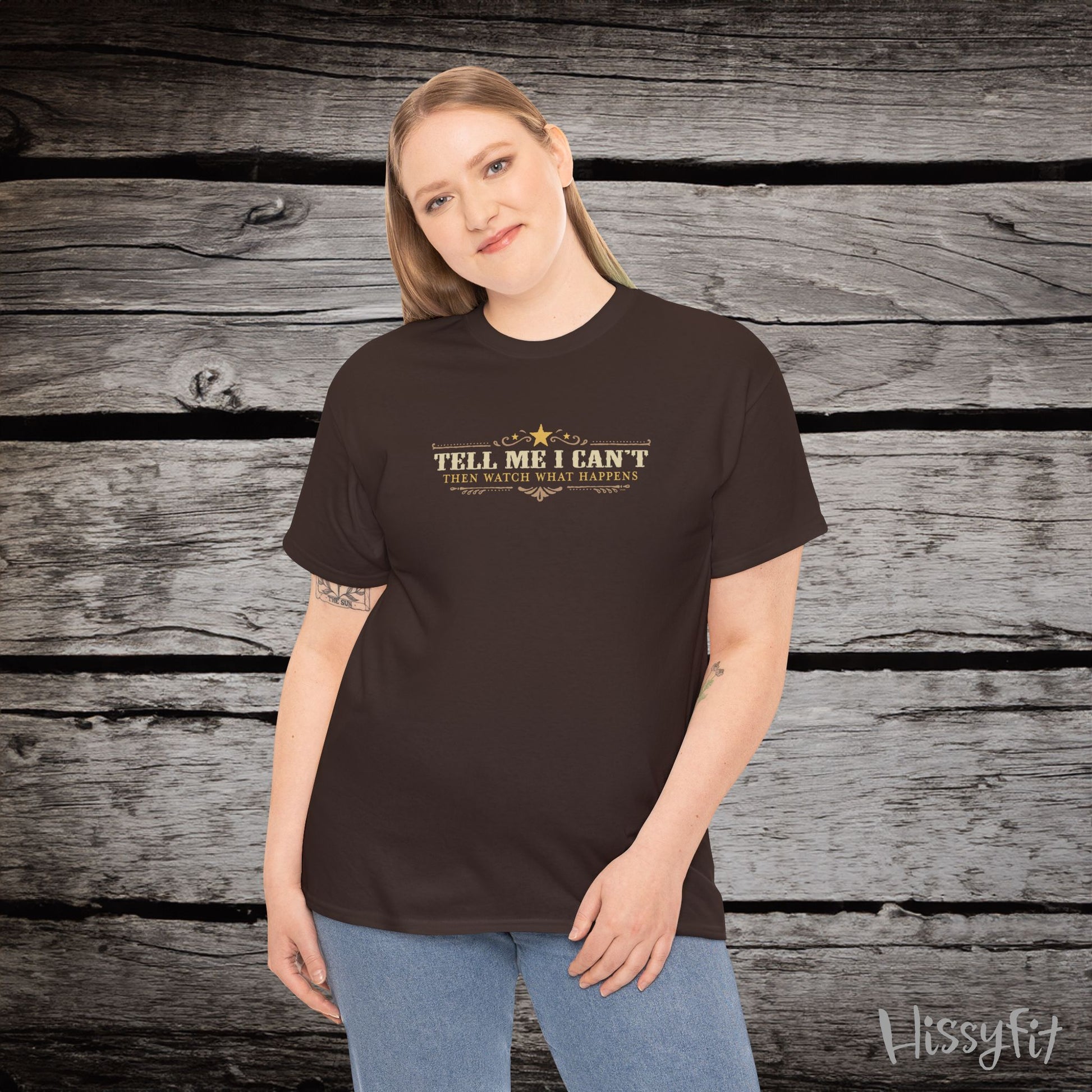 Person wearing a brown t-shirt with text 'Tell me I can't then watch what happens' on a wooden surface, featuring the brand 'Hissyfit'.