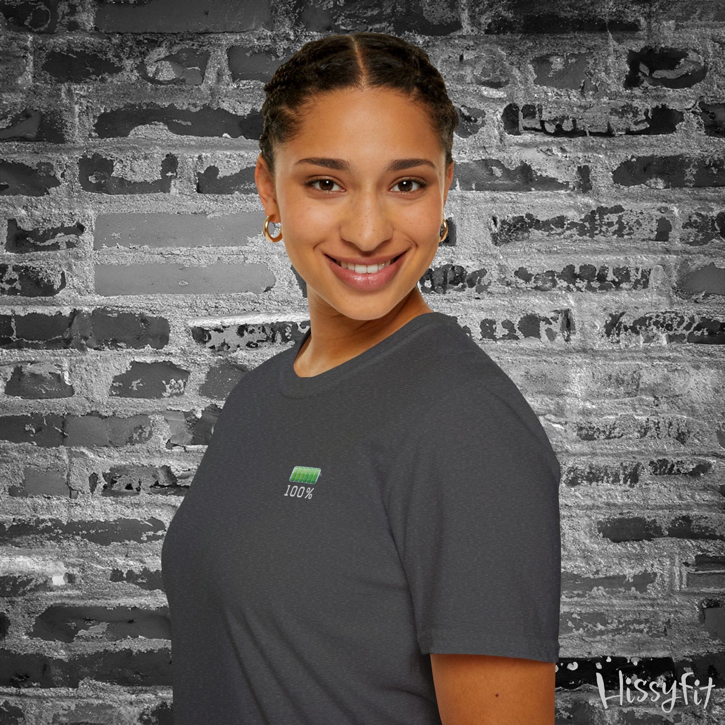 Person wearing a gray t-shirt with a green battery symbol displaying '100 percent charged' on a brick wall background