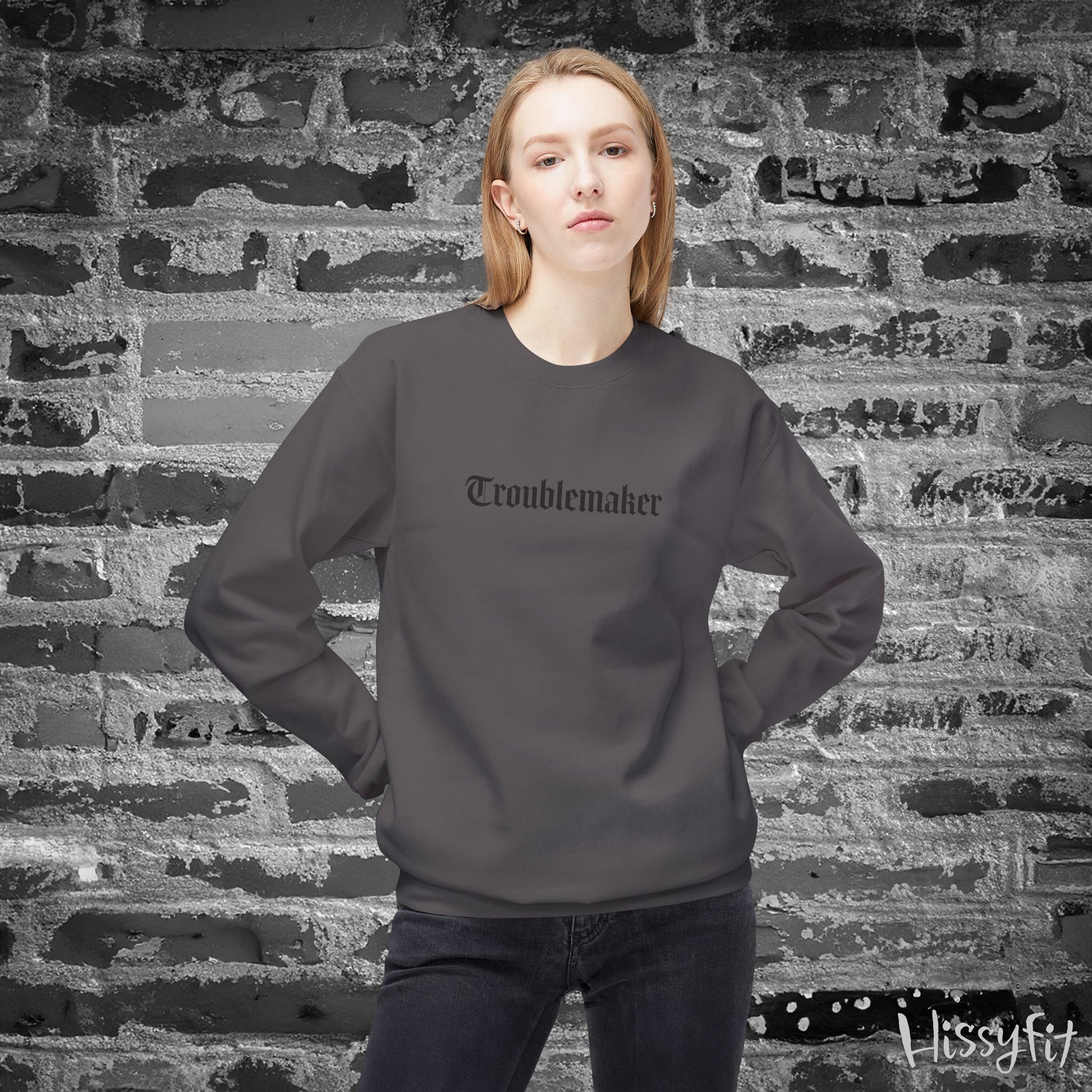 Person wearing a gray sweatshirt with 'Troublemaker' text against a textured brick wall.