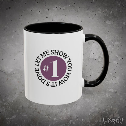 White mug with black interior and handle, featuring text 'Let me show you how it's done' and a number one symbol on a gray background.