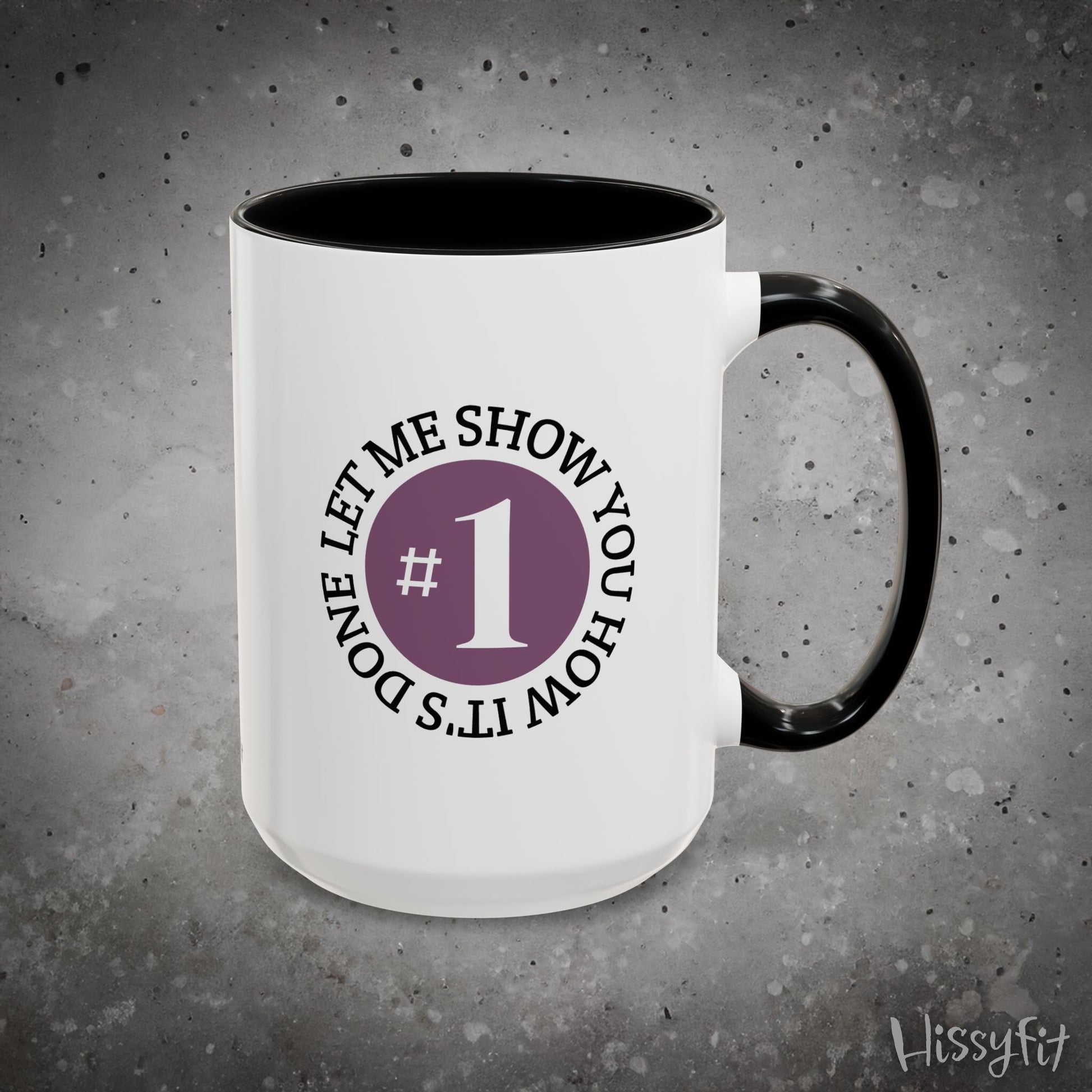 White mug with black interior and handle on a gray background, featuring text 'Let me show you how it's done' and a number one symbol.