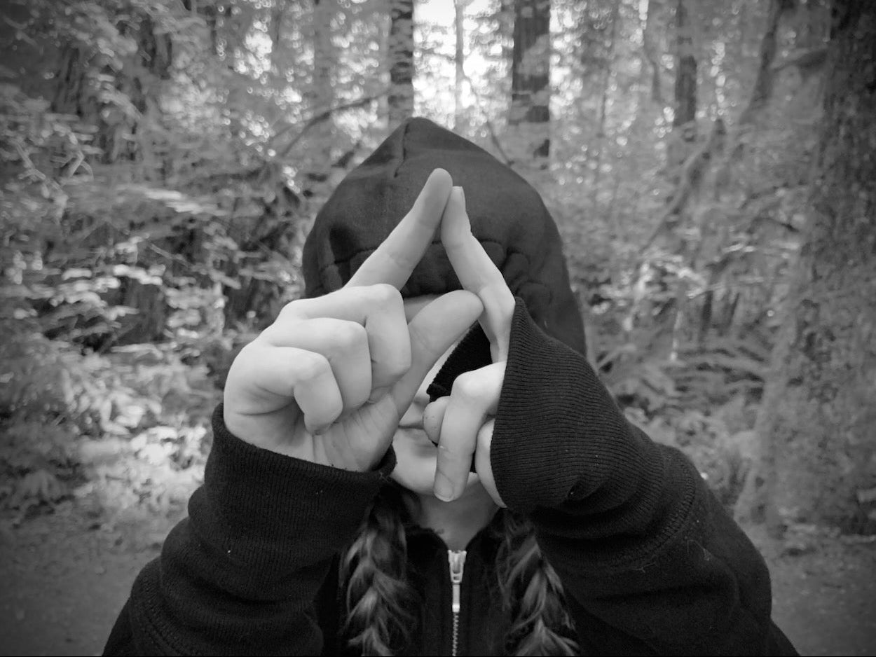 Person in a dark hoodie making a hand gesture in a forest setting