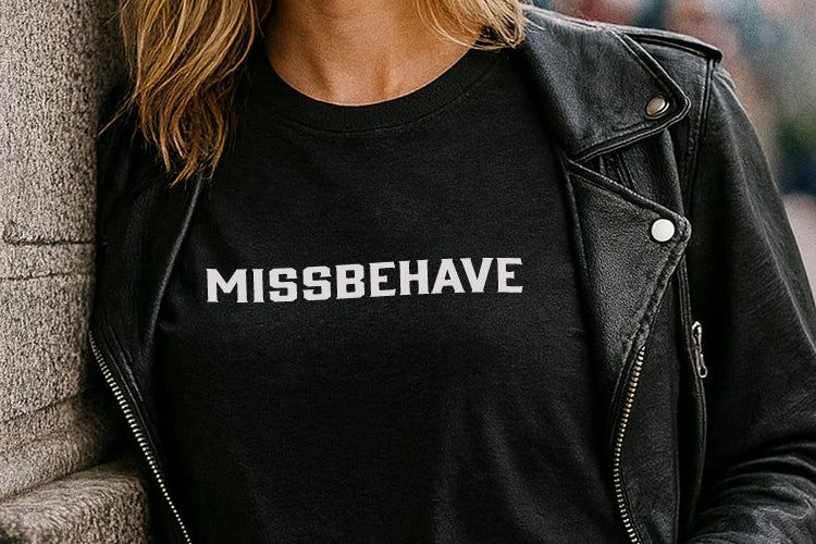 Woman wearing a black 'MISSBEHAVE' t-shirt leaning against a wall in an urban setting.